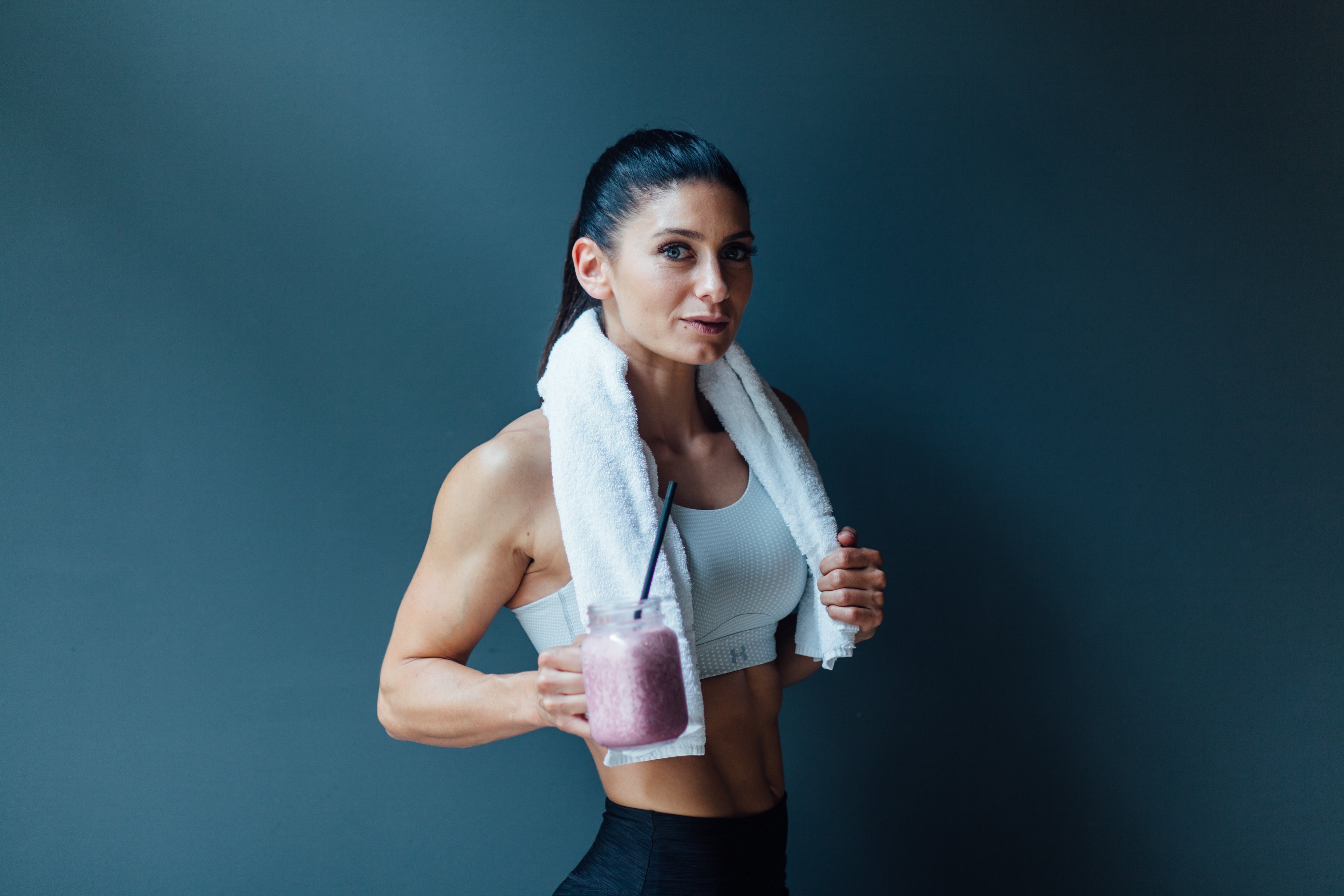 Stefania Lou hält einen lila Smoothie mit Kollagen PURE mit Strohhalm in einem Glas. Sie trägt Sport-BH und Handtuch um den Nacken, vor dunklem Hintergrund.