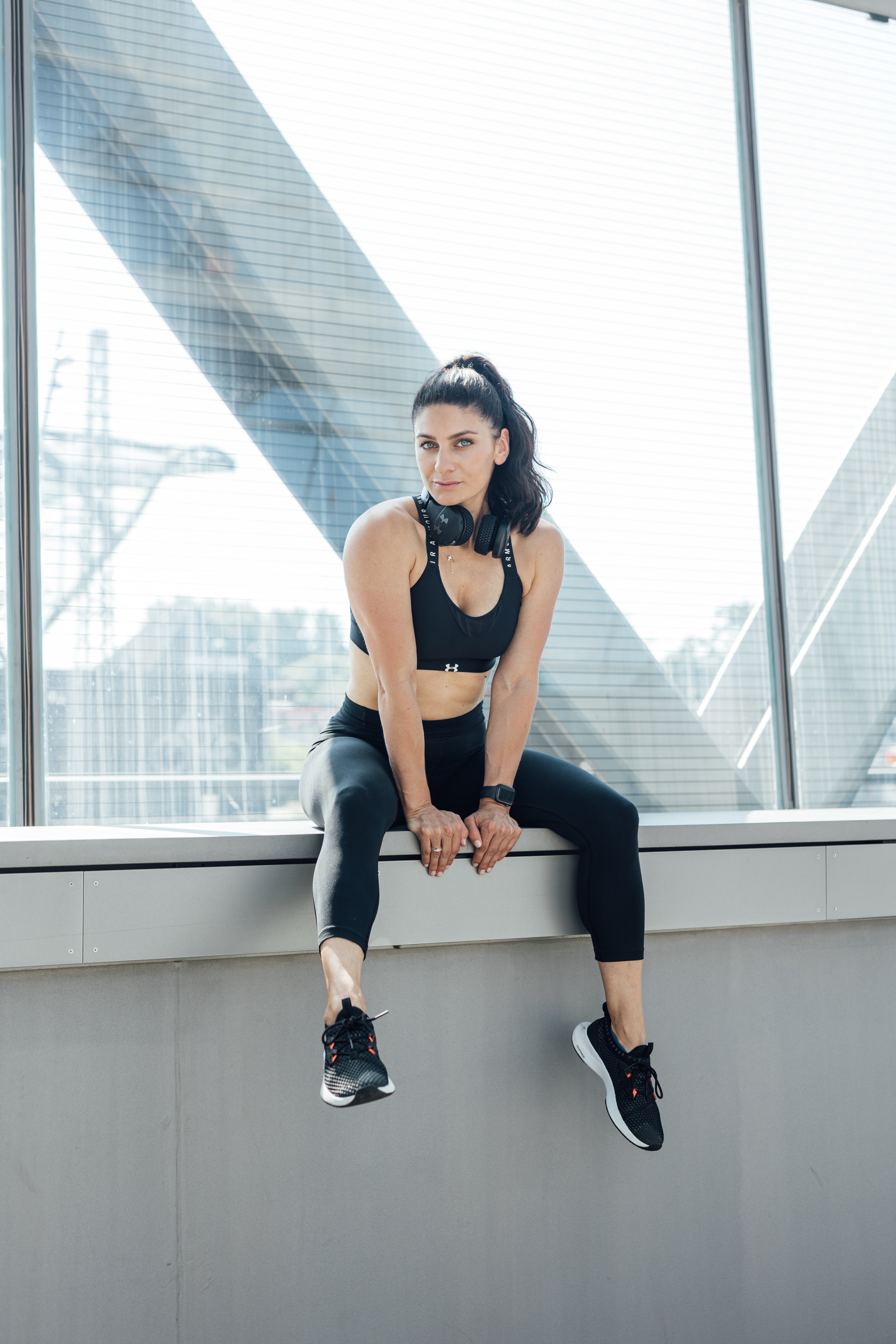 Stefania Lou in schwarzer Sportkleidung sitzt mit Kopfhörern und Smartwatch vor einer Glasfassade.