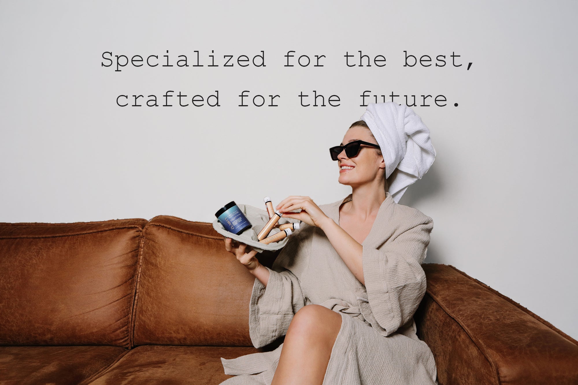 Lachende Frau mit Bademantel, Handtuchturban und Sonnenbrille sitzt auf Sofa und sortiert CLARITY Kollagen Produkte. 